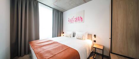 A hotel room with a large bed, a bedside table, a nightstand, a wall-mounted lamp, a painting, and a wardrobe.