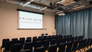 Une salle de conférence avec des rangées de chaises, un écran de projection affichant le texte « ARCHE HOTEL Ozarów Mazowiecki », et un rideau sur le côté droit.