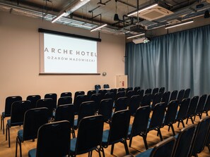 Een vergaderzaal met rijen stoelen, een projectiescherm waarop 'ARCHE HOTEL Ozarów Mazowiecki' staat, en een gordijn aan de rechterkant.