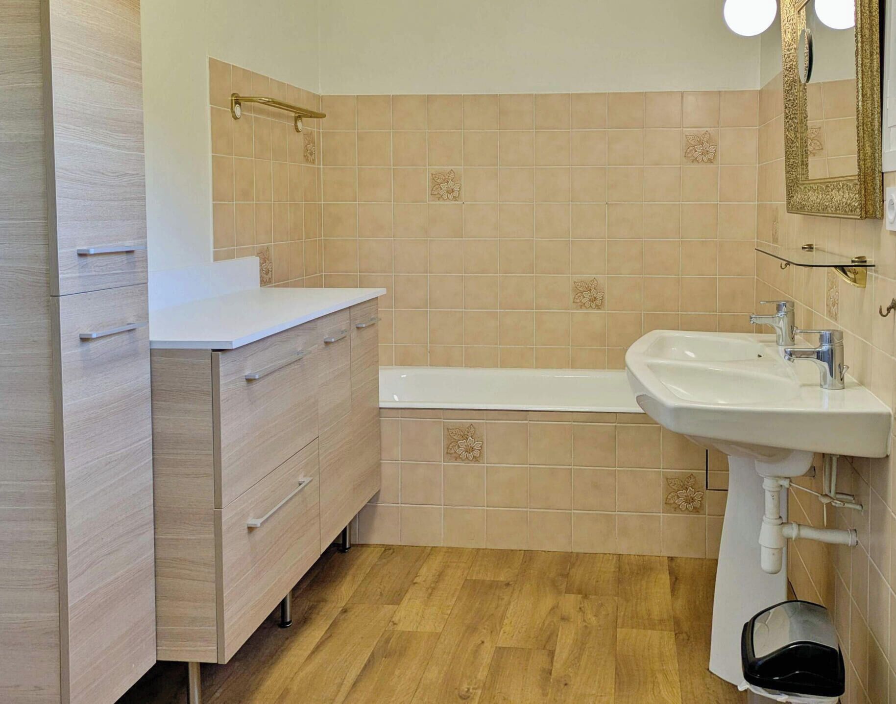 Un baño con piso de madera, azulejos color beige, un lavamanos blanco y un mueble de tocador de madera con cajones.