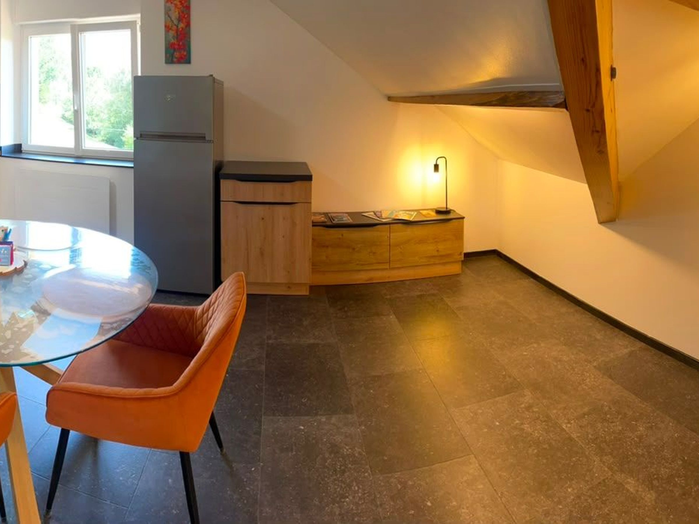 A compact kitchen with a refrigerator, wooden cabinets, a dining table with chairs, and a window with a view of greenery.