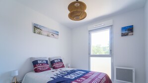 Ein Schlafzimmer mit einem Bett, zwei Kissen, einem Nachttisch, einer Lampe, einem Fenster und einer Wand-Klimaanlage.