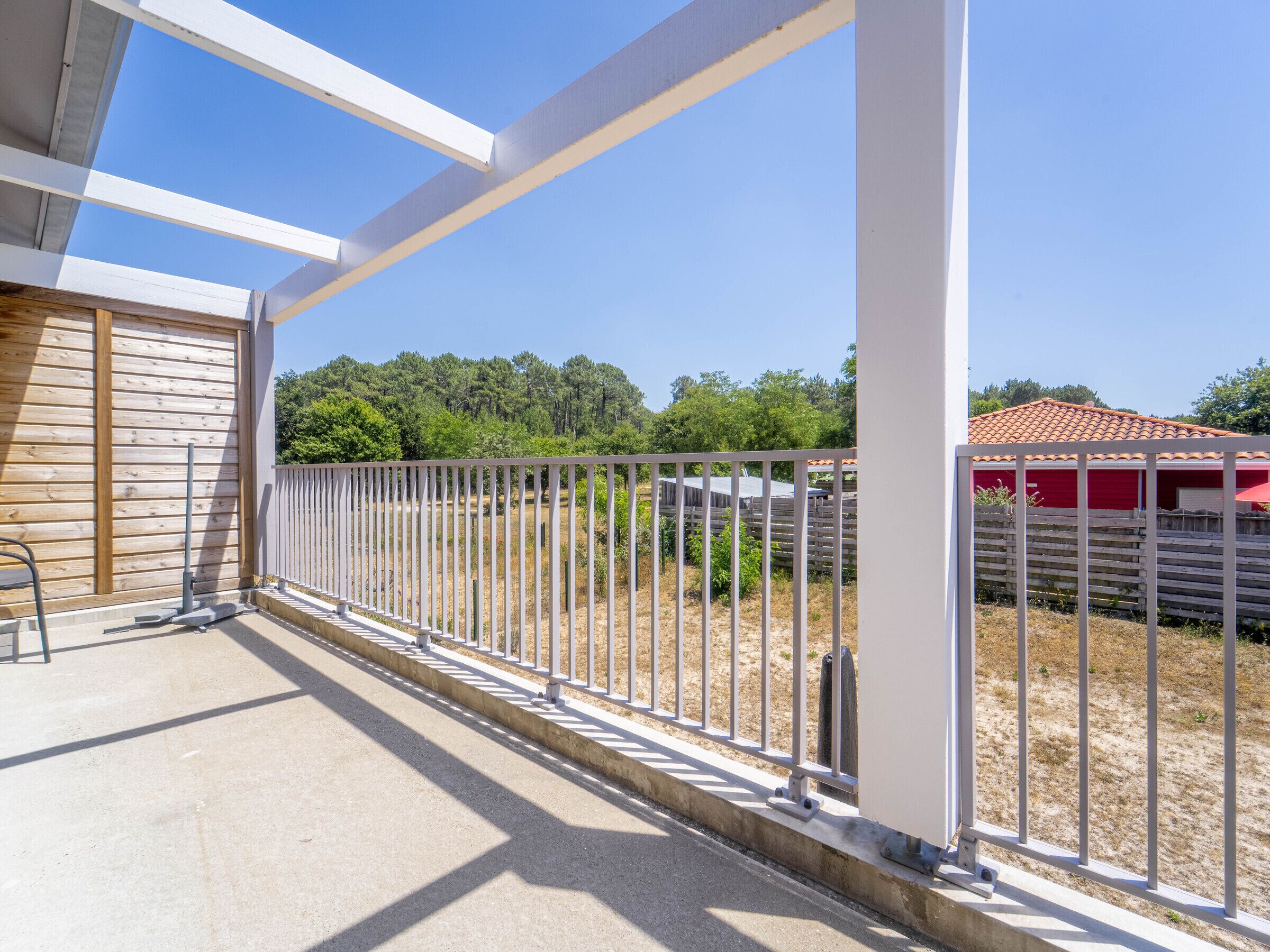 A balcony with a metal railing, a concrete floor, and a view of a fenced area with trees and a red building in the background.