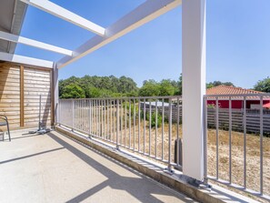 Ein Balkon mit Metallgeländer, einem Betonboden und Blick auf einen eingezäunten Bereich mit Bäumen und einem roten Gebäude im Hintergrund.