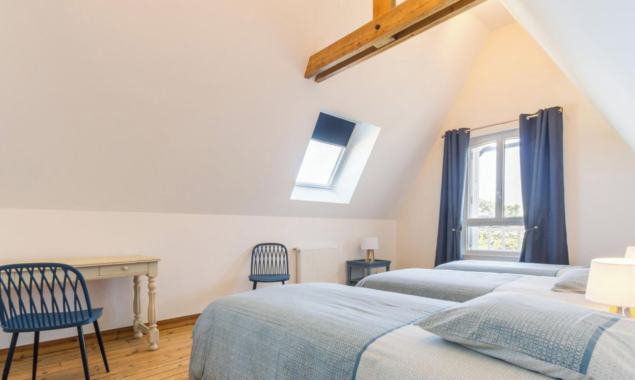 Un dormitorio con techo inclinado, tragaluz, escritorio, silla, cama con dos almohadas y ventana con cortinas.