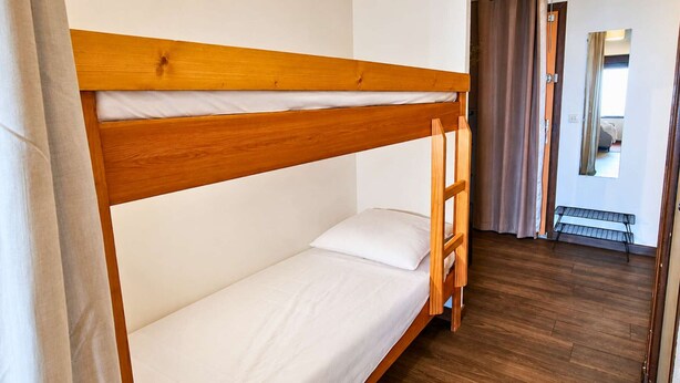 Ein Stockbett mit Holzrahmen und weißer Bettwäsche in einem Zimmer mit Holzboden und einer Tür zu einem weiteren Raum.