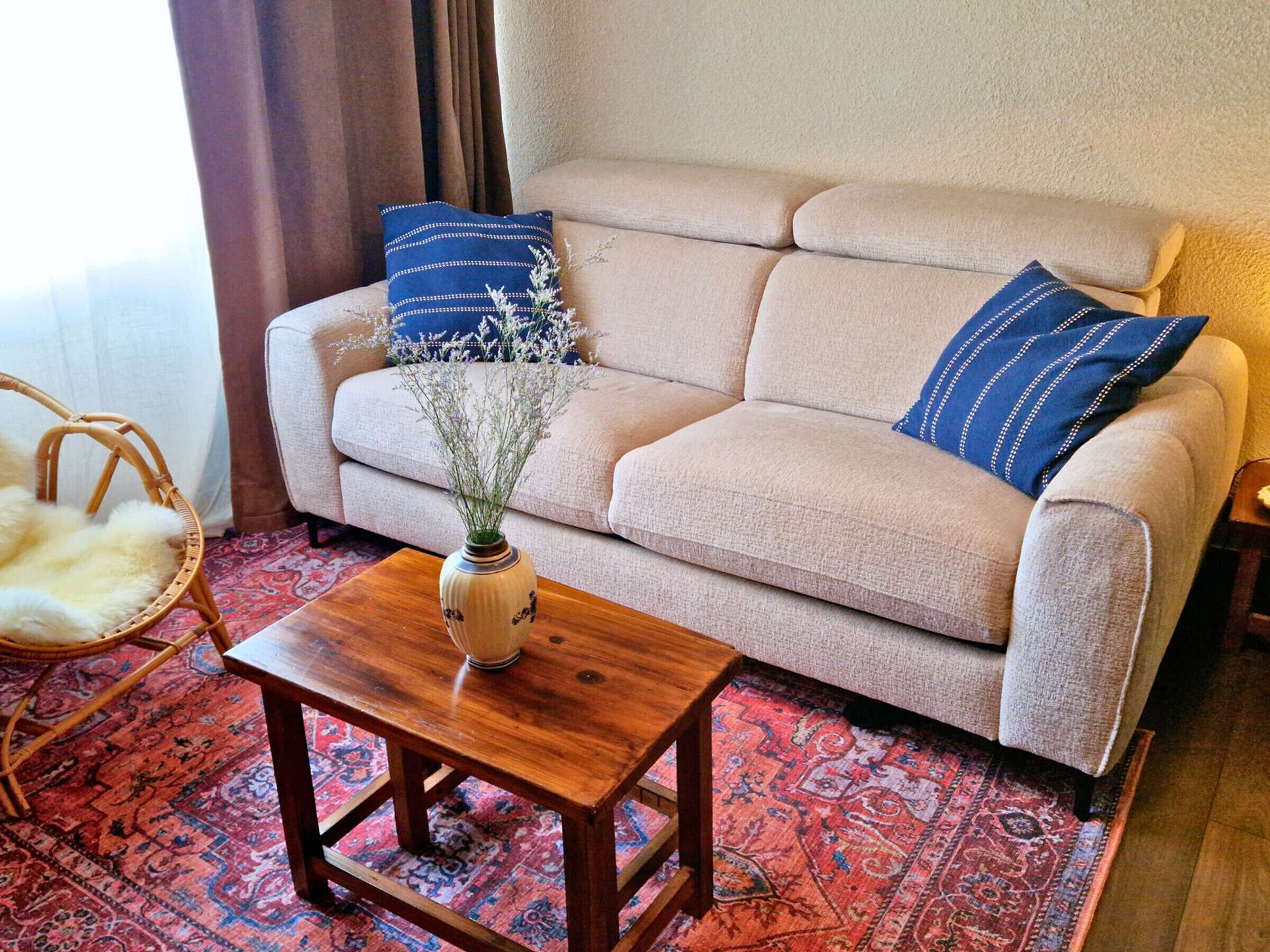 Een woonkamer met een beige bank, twee blauwe kussens, een houten salontafel, een rieten fauteuil en een vloerkleed met dessin.