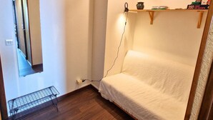 Ein kleiner, schmaler Raum mit einem Einzelbett, einem Spiegel, einem Regal mit Büchern und einem Wasserkocher, sowie einer Steckdose an der Wand.