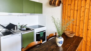 Eine kleine Küche mit Herd, Spüle und grünen Schränken. Ein Holztisch mit Stühlen und eine Vase mit Pflanzen steht bereit.