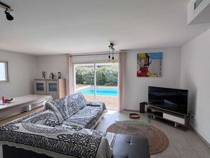 A modern living room with a sofa, a TV, and a glass door leading to a pool. - Villa with pool, WiFi - 10 guests (Moliets-et-Maa)