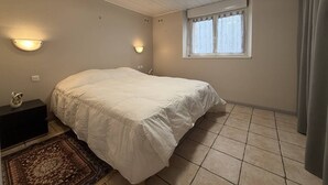 Et soveværelse med seng, et natbord, vægmonteret lampe, et vindue med gardiner og et flisegulv.