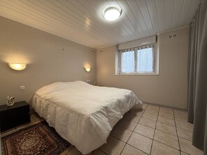 Ein Schlafzimmer mit einem Bett, einem Nachttisch, einer Wandleuchte, einem Fenster mit Vorhängen und einem gefliesten Boden.