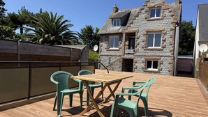 En terrasse med et træbord og fire stole, en bygning af sten i baggrunden og et træhegn.