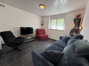 Ein modernes Wohnzimmer mit einem Flachbildfernseher, einem roten Sessel, einem grauen Sofa und einem Fenster mit Blick auf Grünflächen.