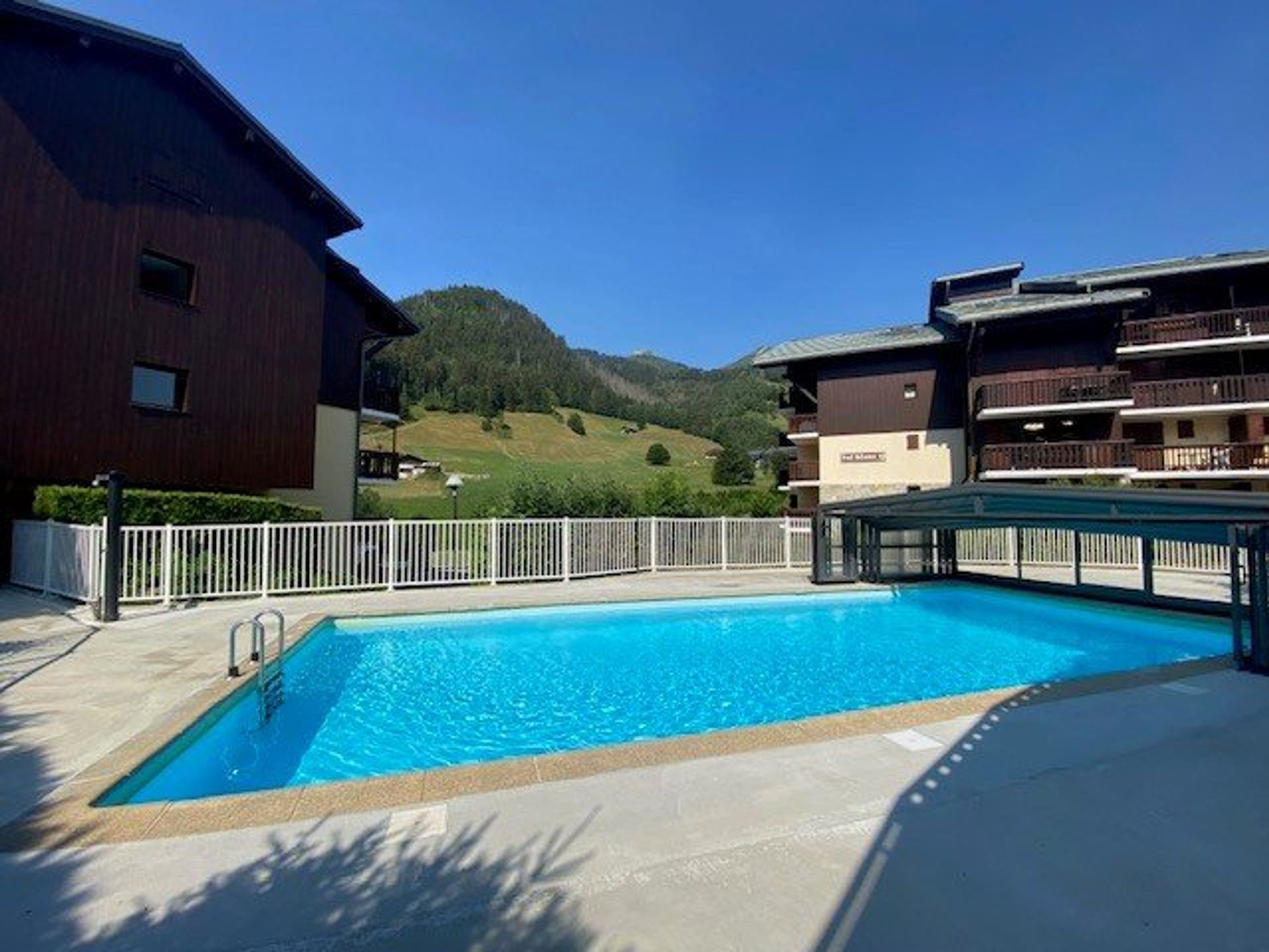 A swimming pool with a white fence, a building with balconies, and a mountainous landscape in the background.
