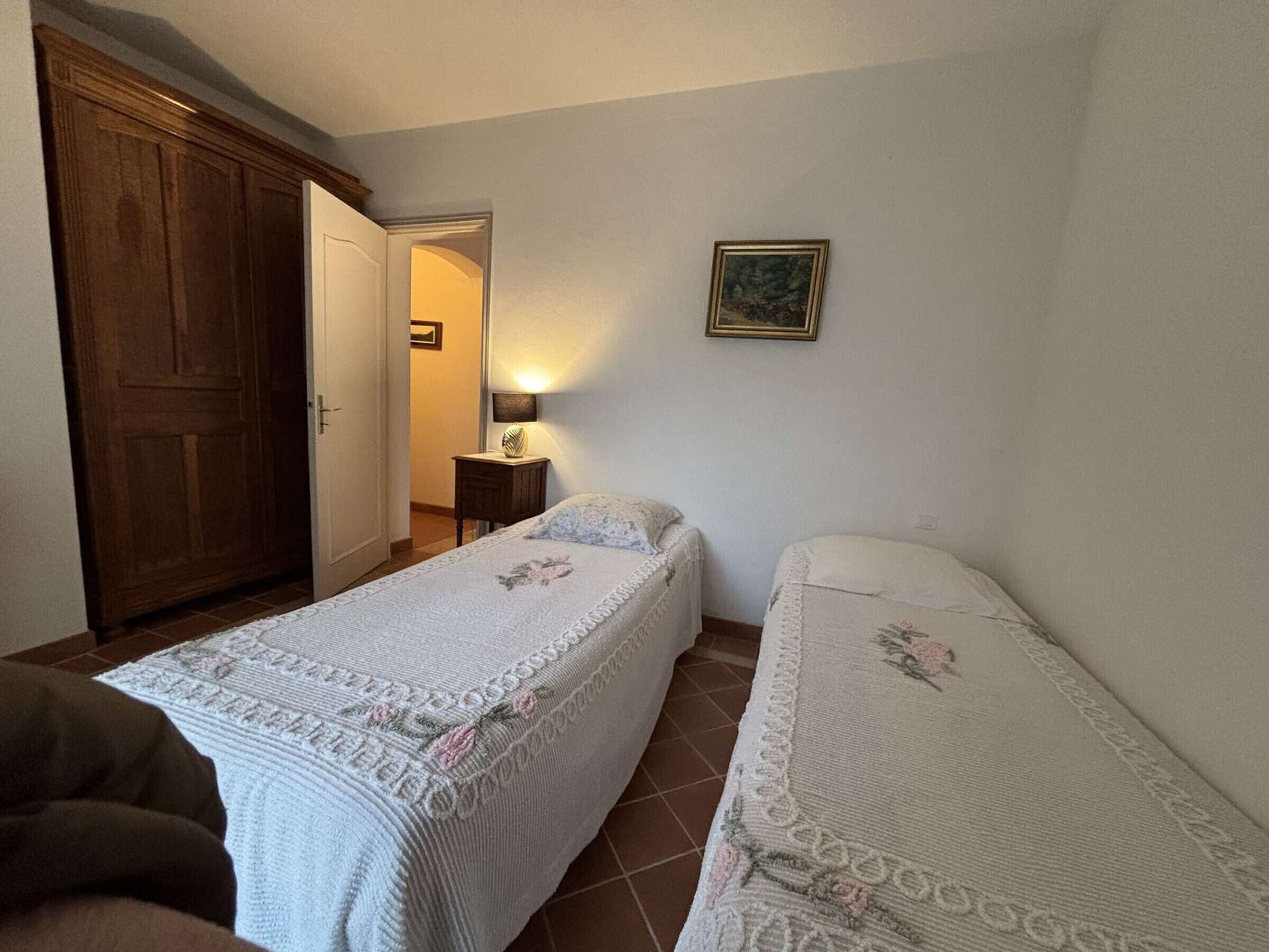 Un dormitorio con dos camas individuales, un armario de madera, una mesita de noche pequeña con una lámpara y un cuadro en la pared.