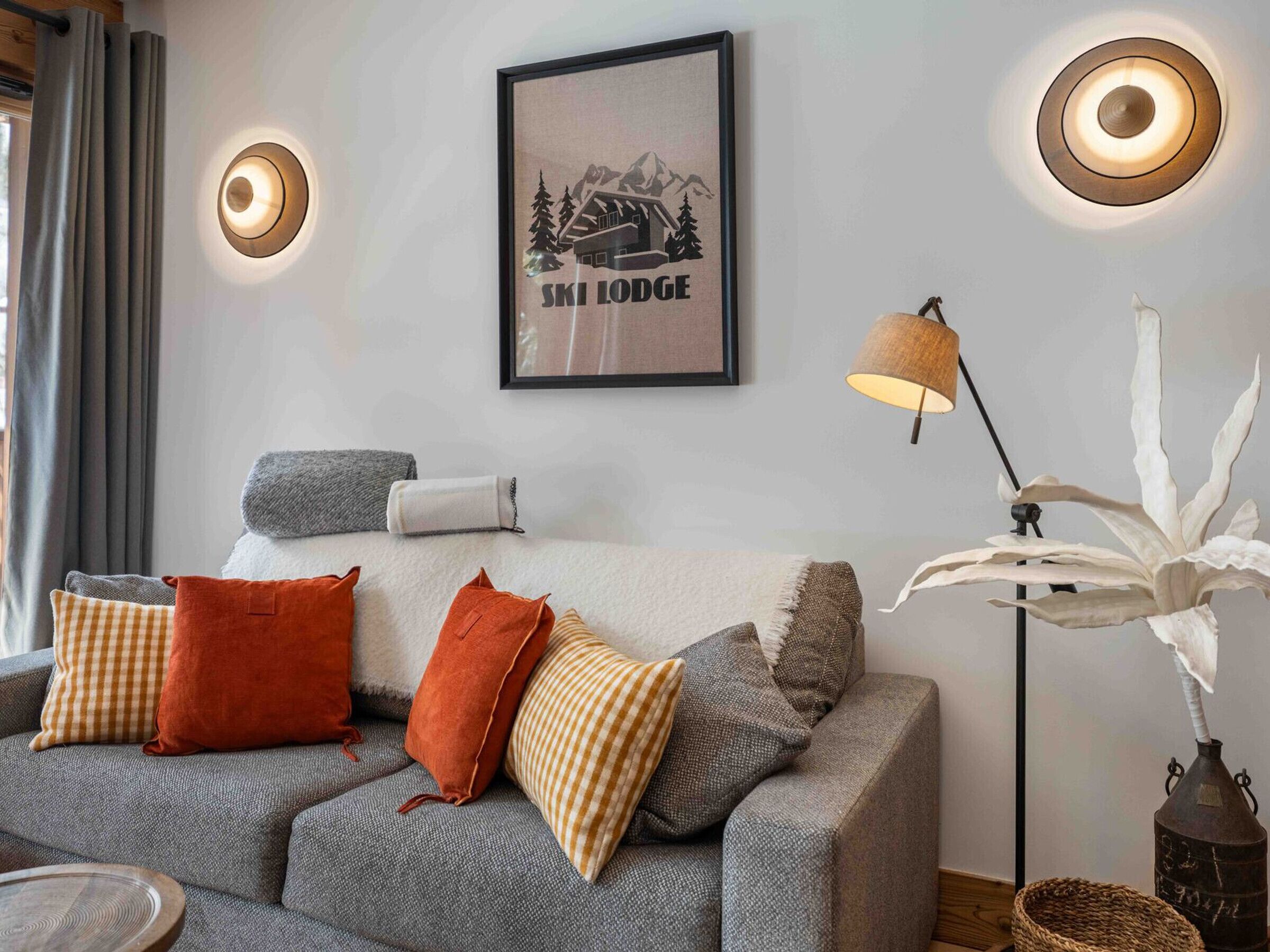 Un salon moderne avec un canapé gris, divers coussins, un tableau encadré représentant un chalet de ski et deux lampes fixées au mur.