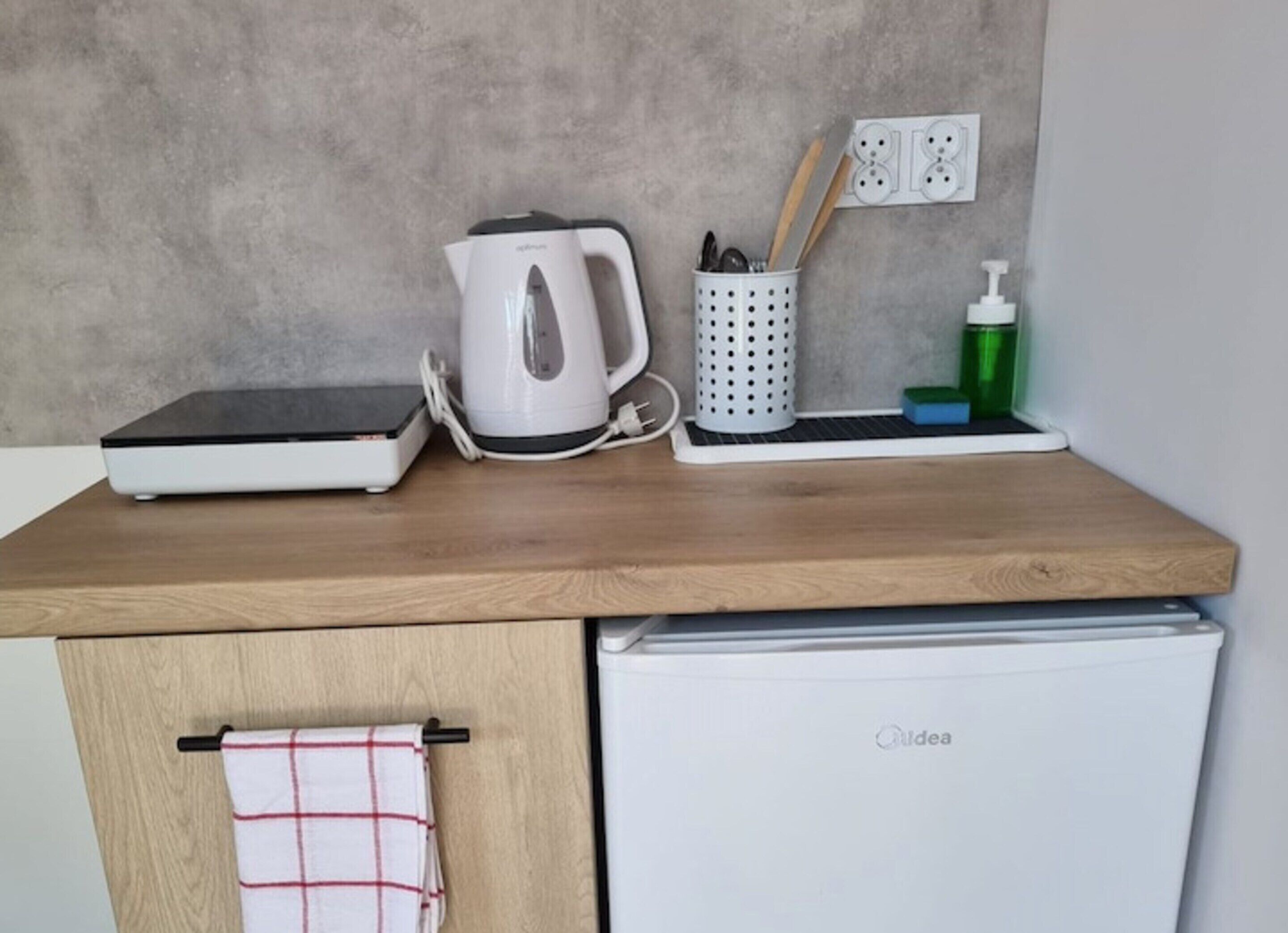Een aanrecht met een koffiezetapparaat, een waterkoker, een container met bestek, een zeepdispenser en een kleine koelkast.