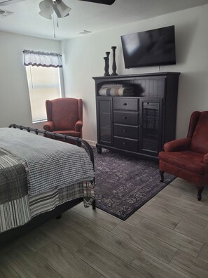 A bedroom with a bed, two armchairs, a dark wooden dresser, a flat-screen TV, and a window with blinds. - 3 bedroom Oasis, Steps from Glendale Stadium (Phoenix)