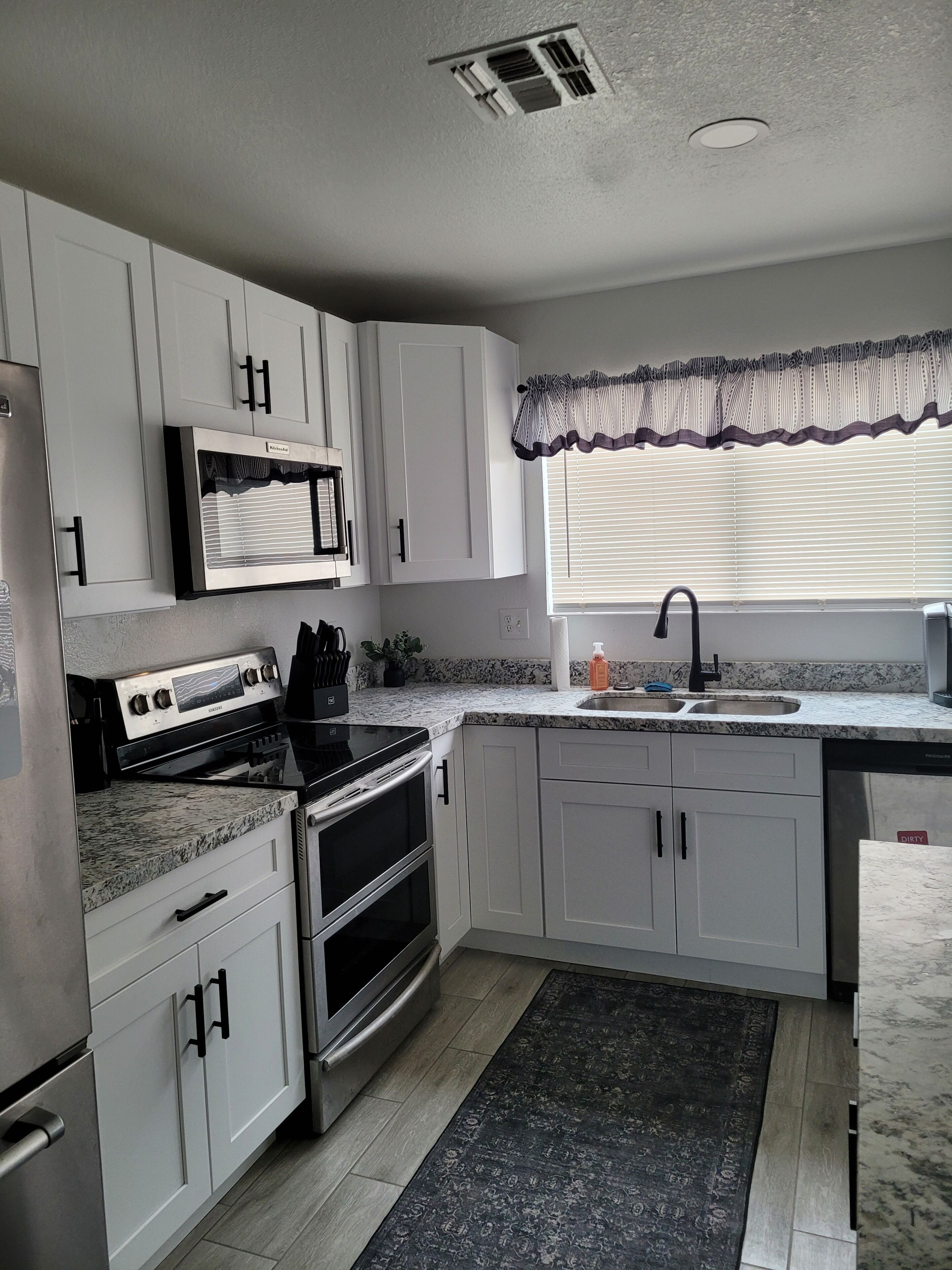 A kitchen with white cabinets, a microwave, an oven, a sink, and a window with blinds.