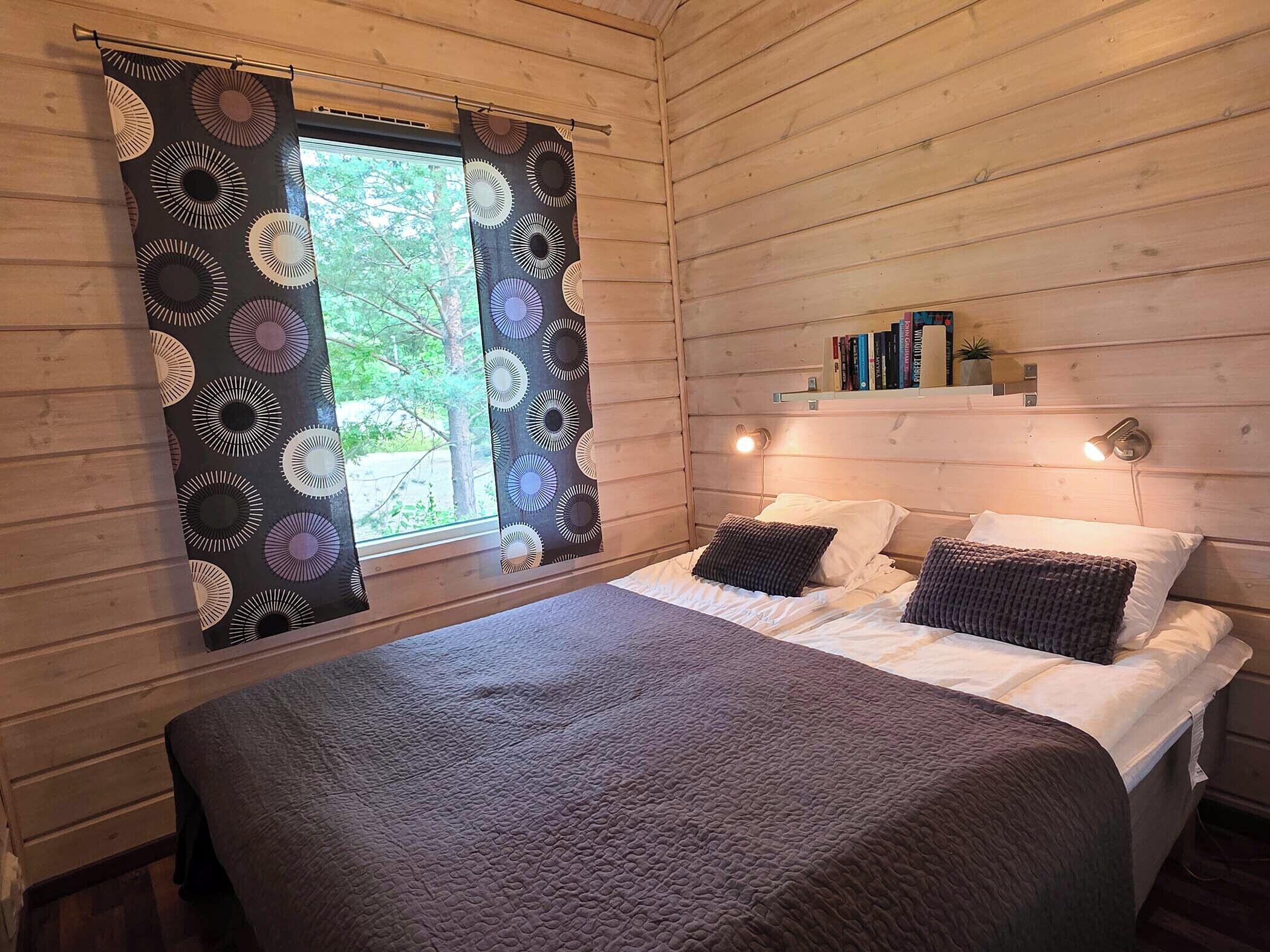 A wooden cabin bedroom with a bed, pillows, a window with curtains, and a shelf with books.
