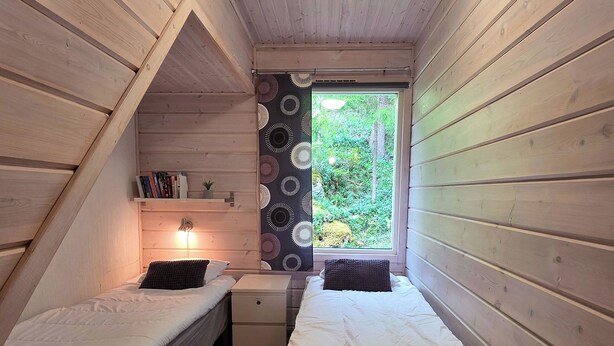 A small, cozy bedroom with two single beds, a nightstand, a shelf with books, and a window with a view of greenery. - Villa Chalet, Himos Länsihuippu (Jämsä)
