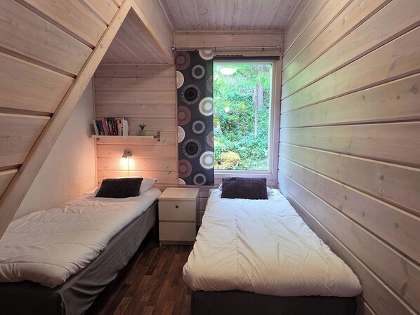 A small, cozy bedroom with two single beds, a nightstand, a shelf with books, and a window with a view of greenery.