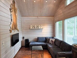 A cozy cabin interior with a wooden ceiling, a mounted television, a sofa, and a window with blinds.