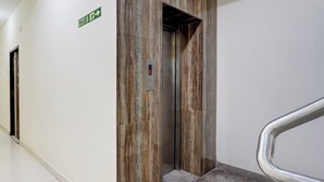 Un pasillo con un letrero de salida de incendios, un ascensor con pantalla digital y un pasamanos de metal.