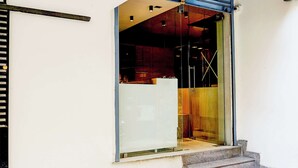 Entrada de un edificio con puerta de vidrio, escalera metálica que sube y un interior visible con una pared roja y un objeto blanco.