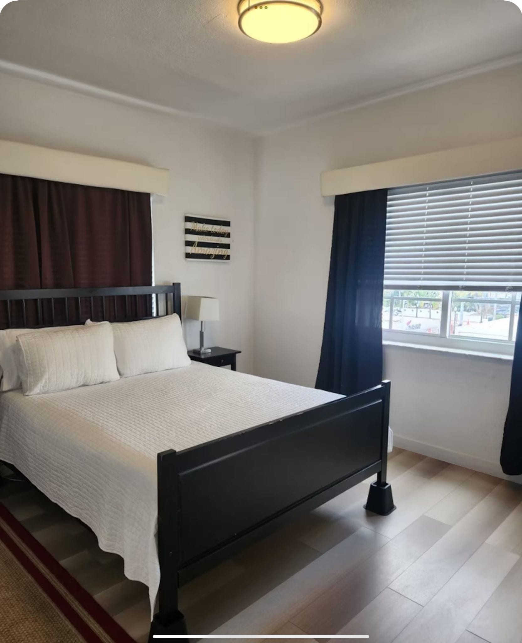 Ein Hotelzimmer mit einem Bett, einem Nachttisch, einer Lampe, einem Fenster mit Vorhängen und einer Wanddekoration.