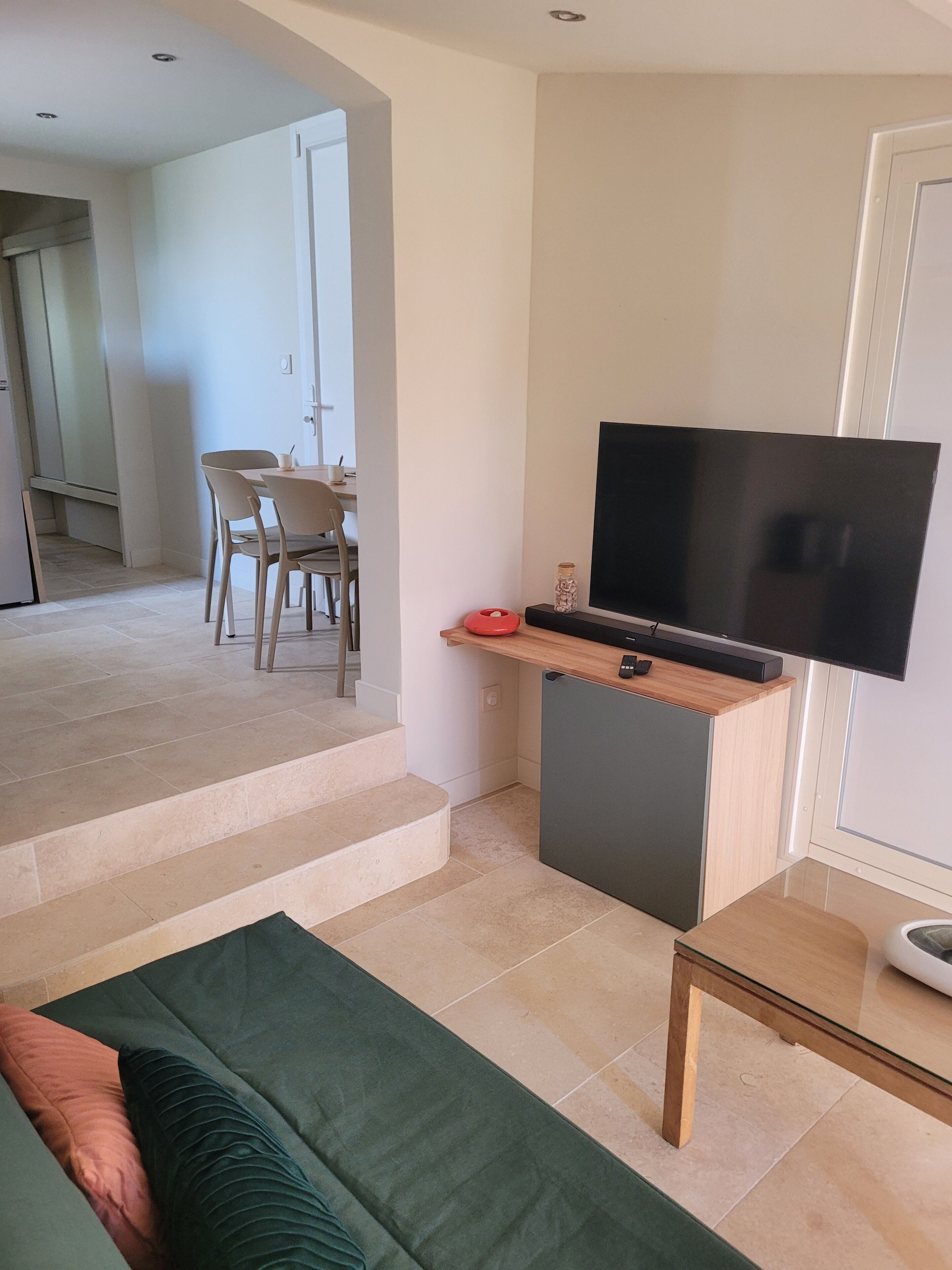 A modern living room with a sofa, a television on a stand, and a dining area in the background.