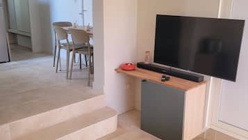 A modern living room with a sofa, a television on a stand, and a dining area in the background.