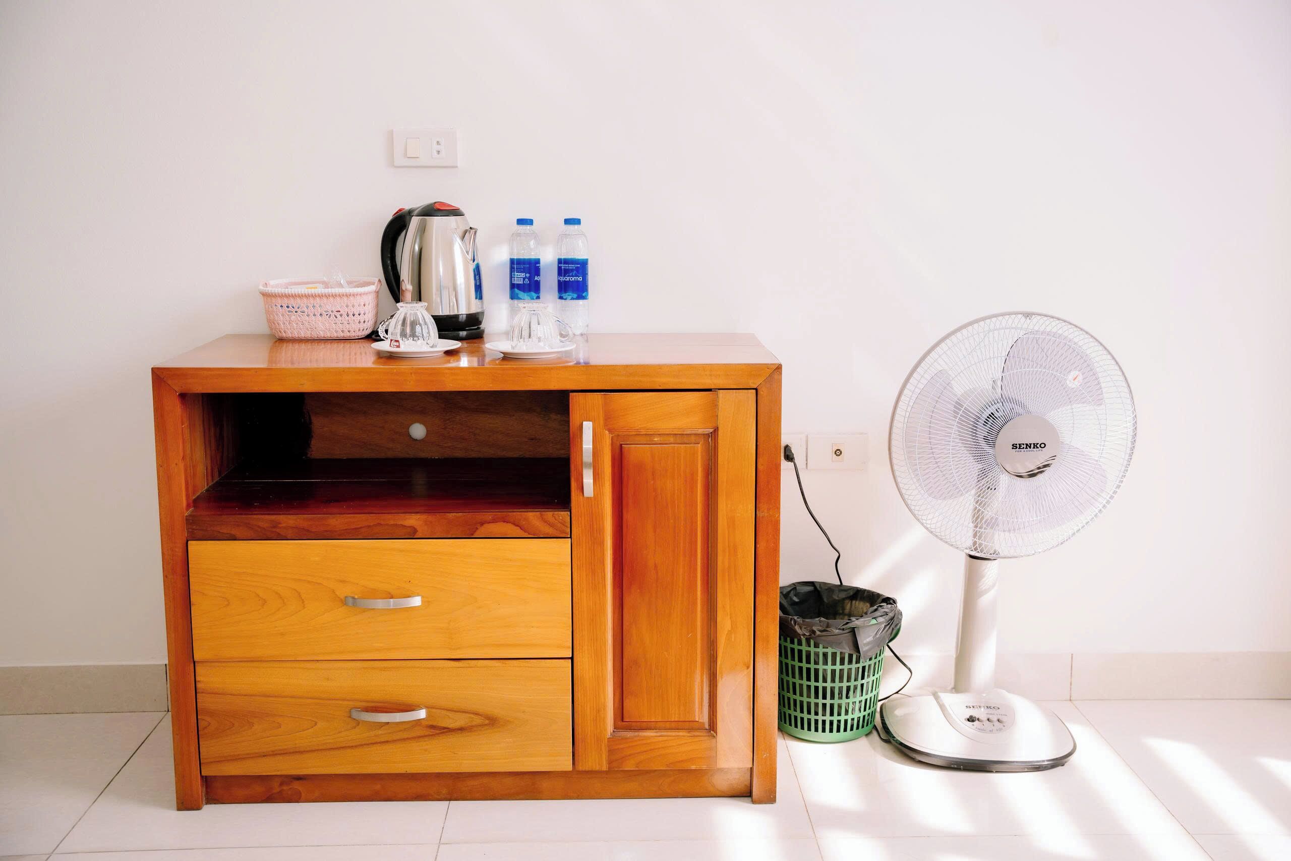 Un mueble de madera con cajones, una tetera, botellas de agua y un ventilador de pie.