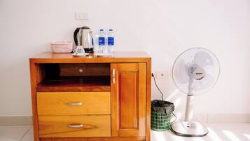 Un mueble de madera con cajones, una tetera, botellas de agua y un ventilador de pie.