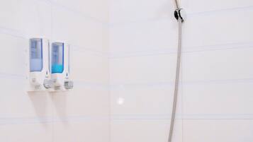 Une douche équipée d’un pommeau de douche à main et d’un robinet mural. Deux distributeurs de savon sont fixés au mur.