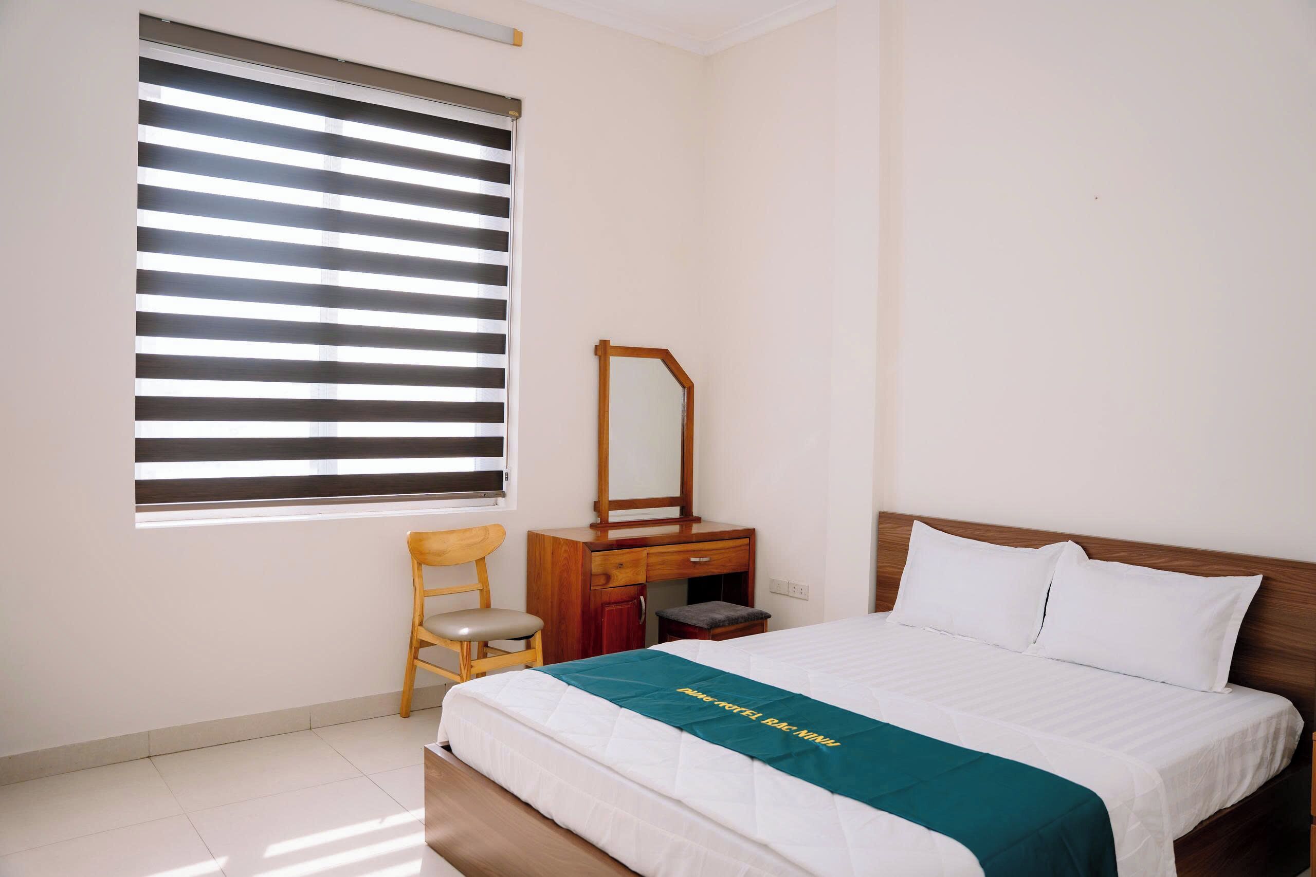 Une chambre d’hôtel avec un lit, une table de chevet en bois, un miroir, une chaise et une fenêtre avec des stores.