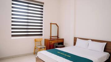 Habitación de hotel con cama, mesita de noche de madera, espejo, silla y ventana con persianas.