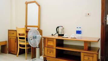 A small, neatly arranged room with a wooden desk, a chair, a mirror, a fan, a kettle, and two bottles of water.