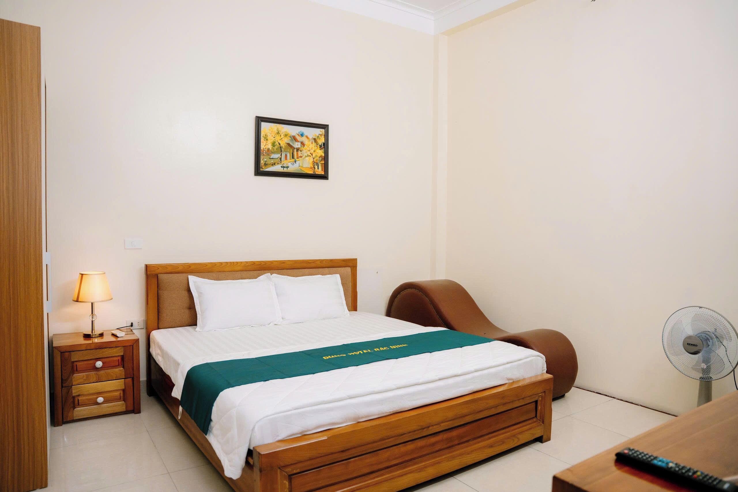 Une chambre d’hôtel avec un lit en bois, une table de chevet avec une lampe, une chaise, un bureau avec un ventilateur et un tableau accroché au mur.