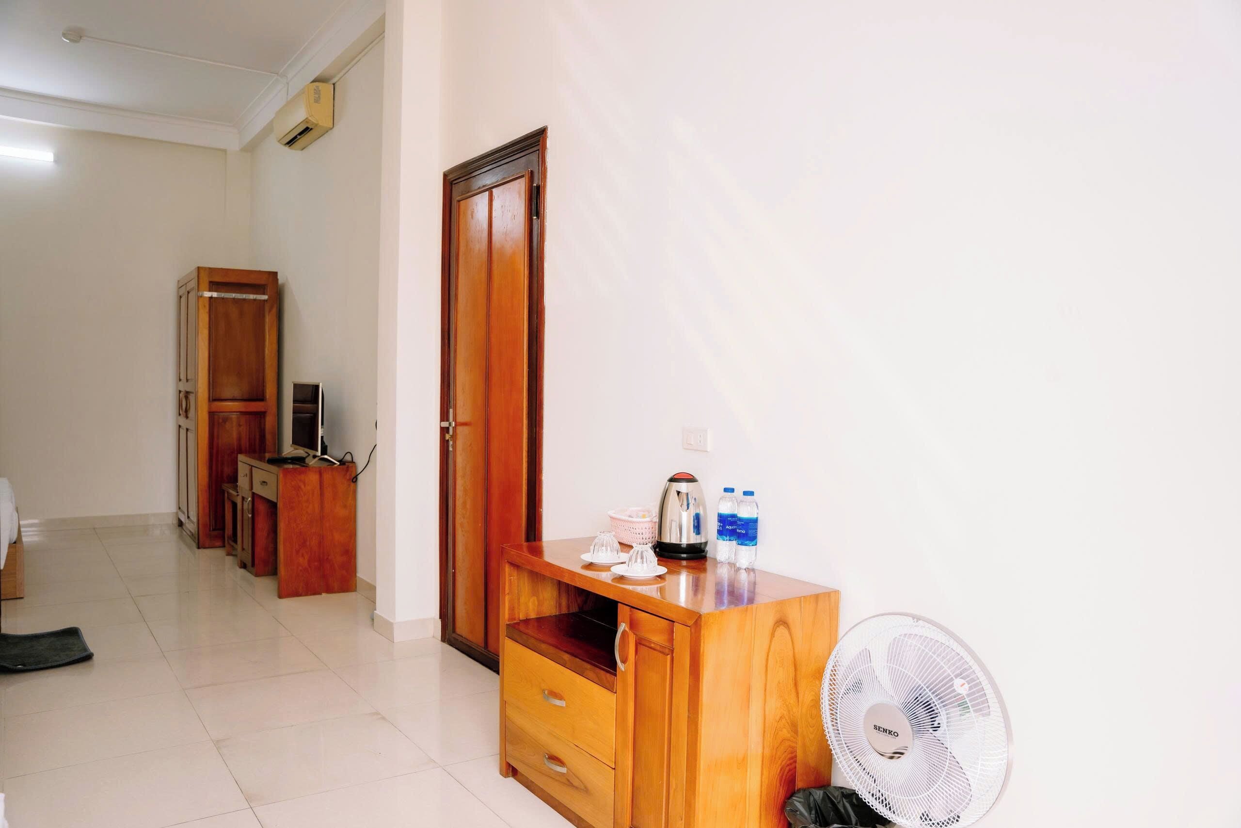 A hotel room with a wooden wardrobe, a desk with a kettle, a fan, and a television.