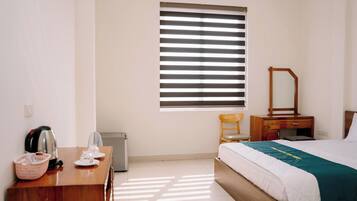 Habitación de hotel con cama, escritorio con hervidor, silla, espejo y ventana con persianas.