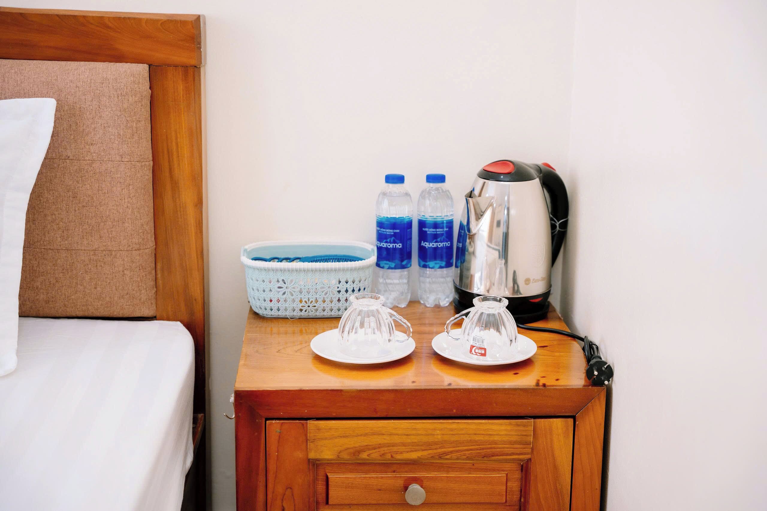 A wooden bedside table with a white kettle, two water bottles, a glass carafe, and a white basket.