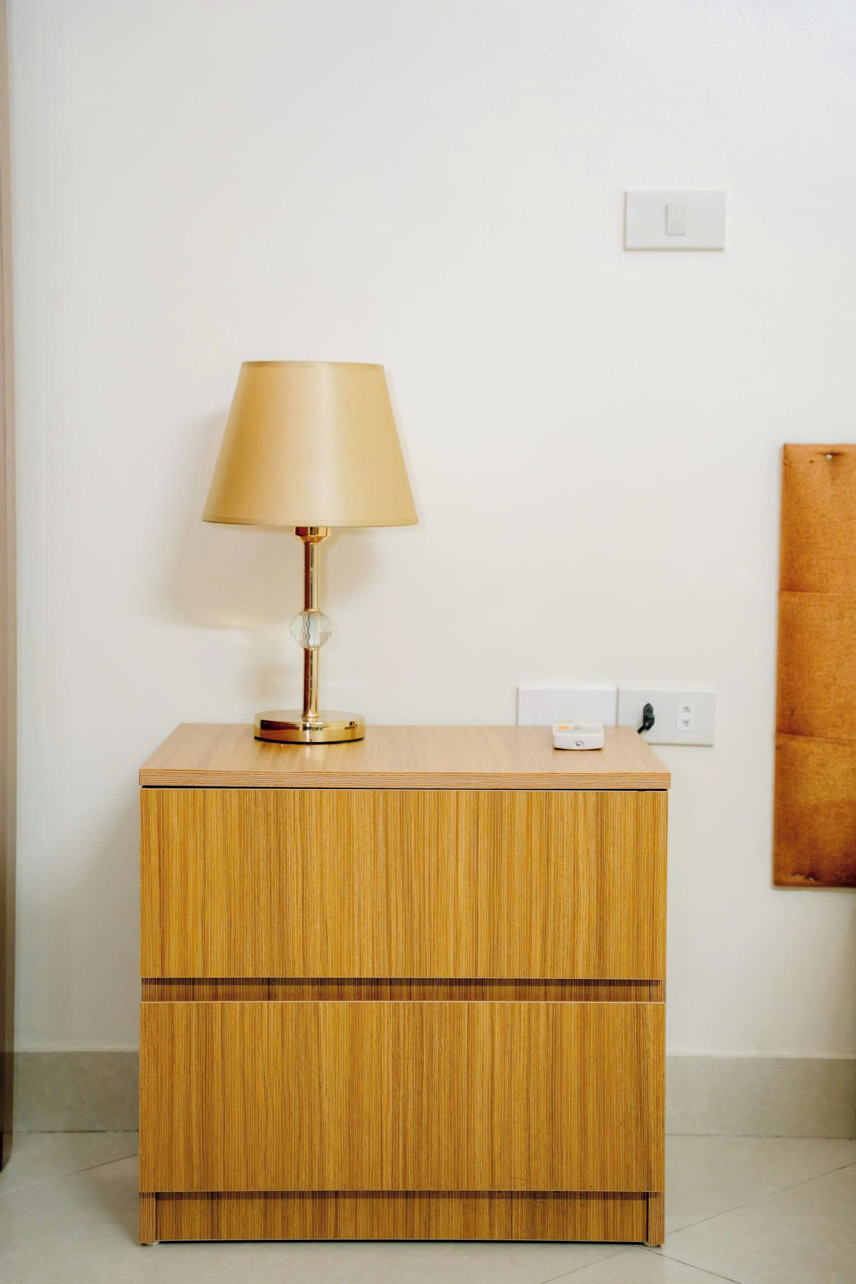 A wooden bedside table with a lamp and a power outlet on the wall.
