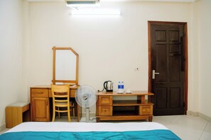 A bedroom with a bed, a desk with a chair, a mirror, a fan, a kettle, and a door. - Lotus Hotel 1 (Hap Linh)