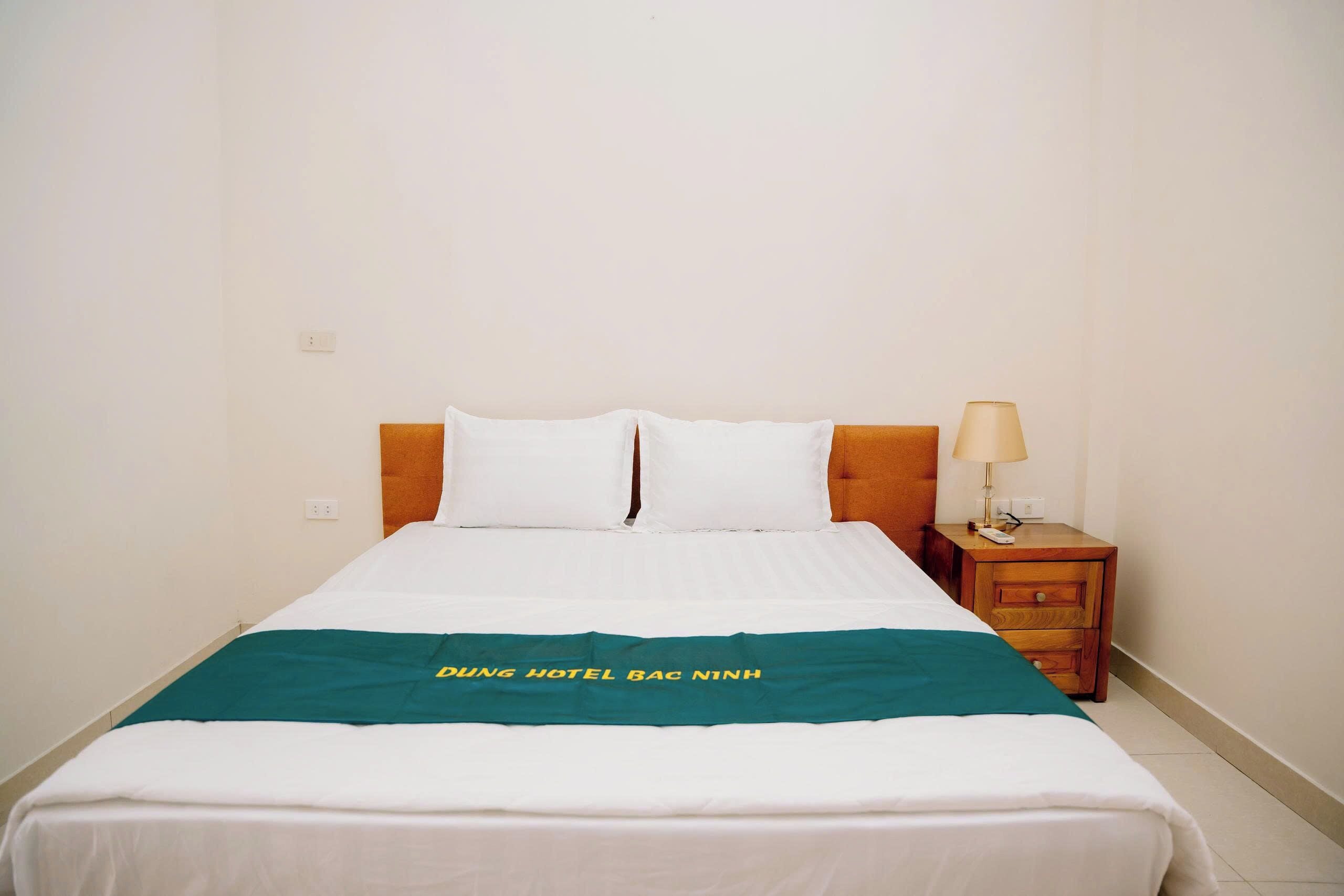 Una cama individual con ropa blanca de cama y una manta verde con texto, una mesita de noche de madera con una lámpara, y una pared blanca lisa.