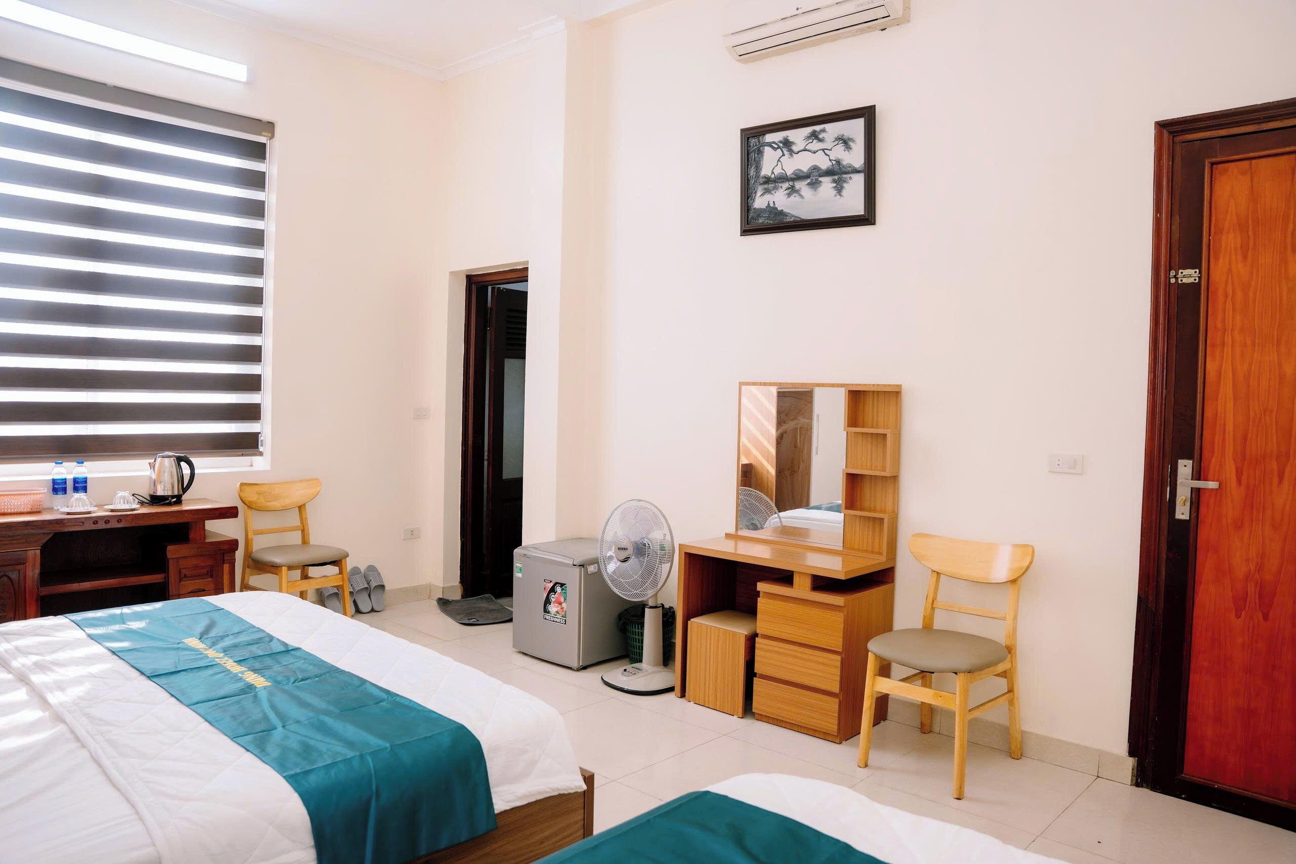 Quarto de hotel com duas camas, uma escrivaninha, uma cadeira, um ventilador, um guarda-roupa e um quadro na parede.