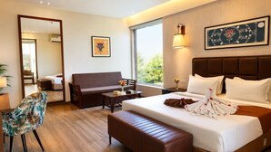 A hotel room with a large bed, a sofa, a small table, a chair, and a window with a view of greenery.
