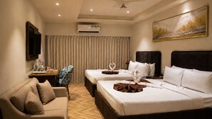 A hotel room with two beds, a sofa, a desk with a chair, a ceiling fan, an air conditioning unit, and a painting on the wall.
