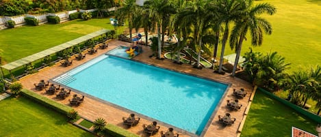 Piscina rectangular con cubierta de madera, rodeada de palmeras y césped.